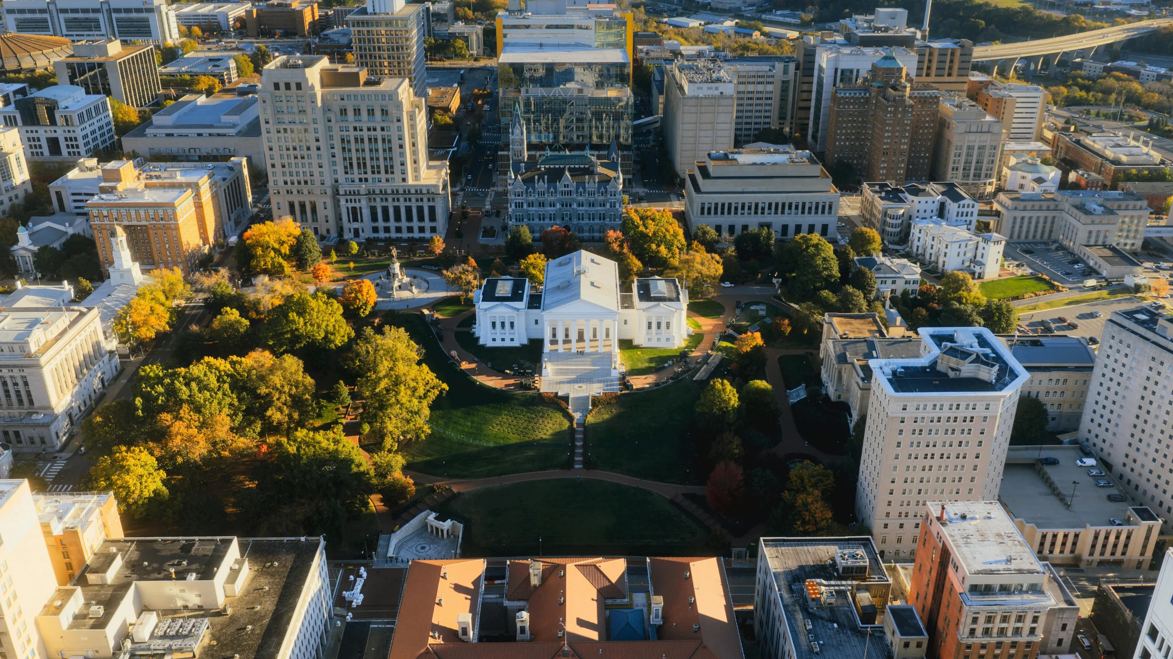 Aerial view of a white capitol-style government building surrounded by trees and downtown office blocks.