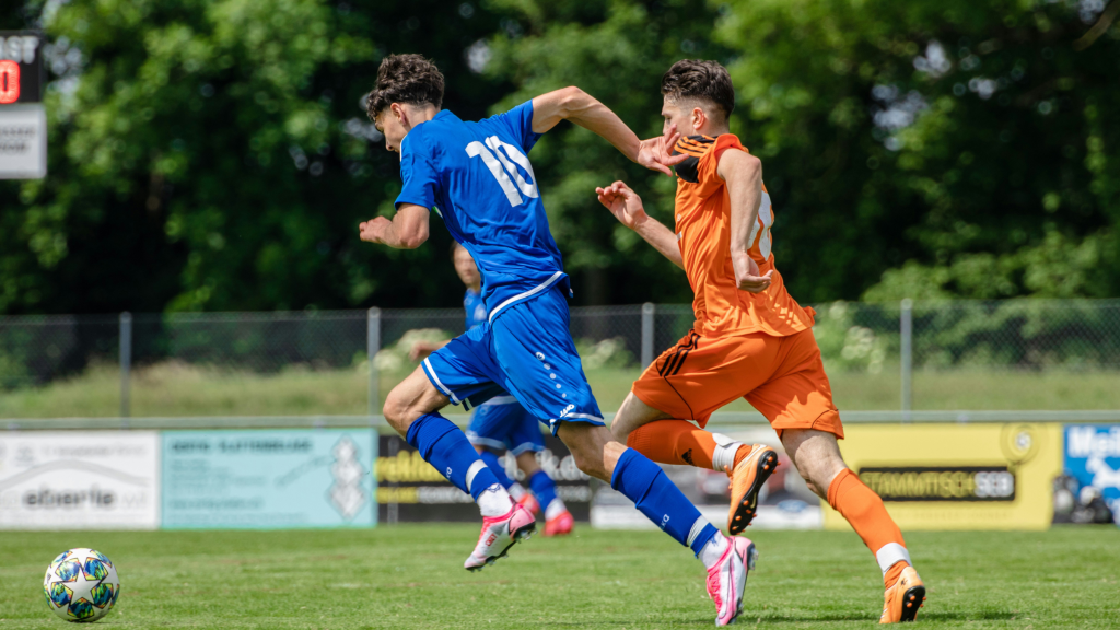 Two soccer players in blue and orange kits sprint toward the ball during a match.