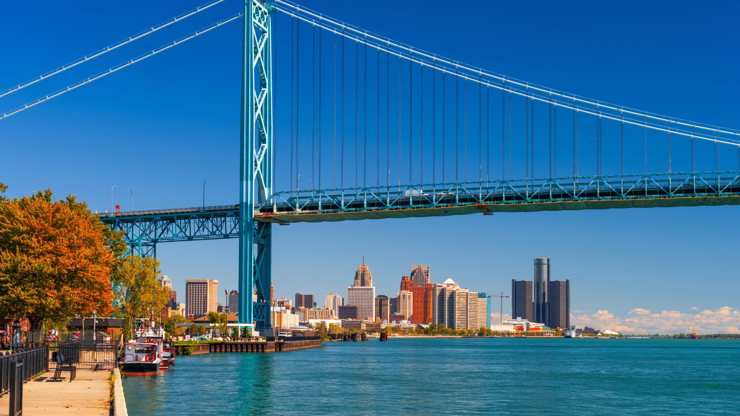 Detroit skyline seen from the waterfront beneath a large suspension bridge on a clear day.