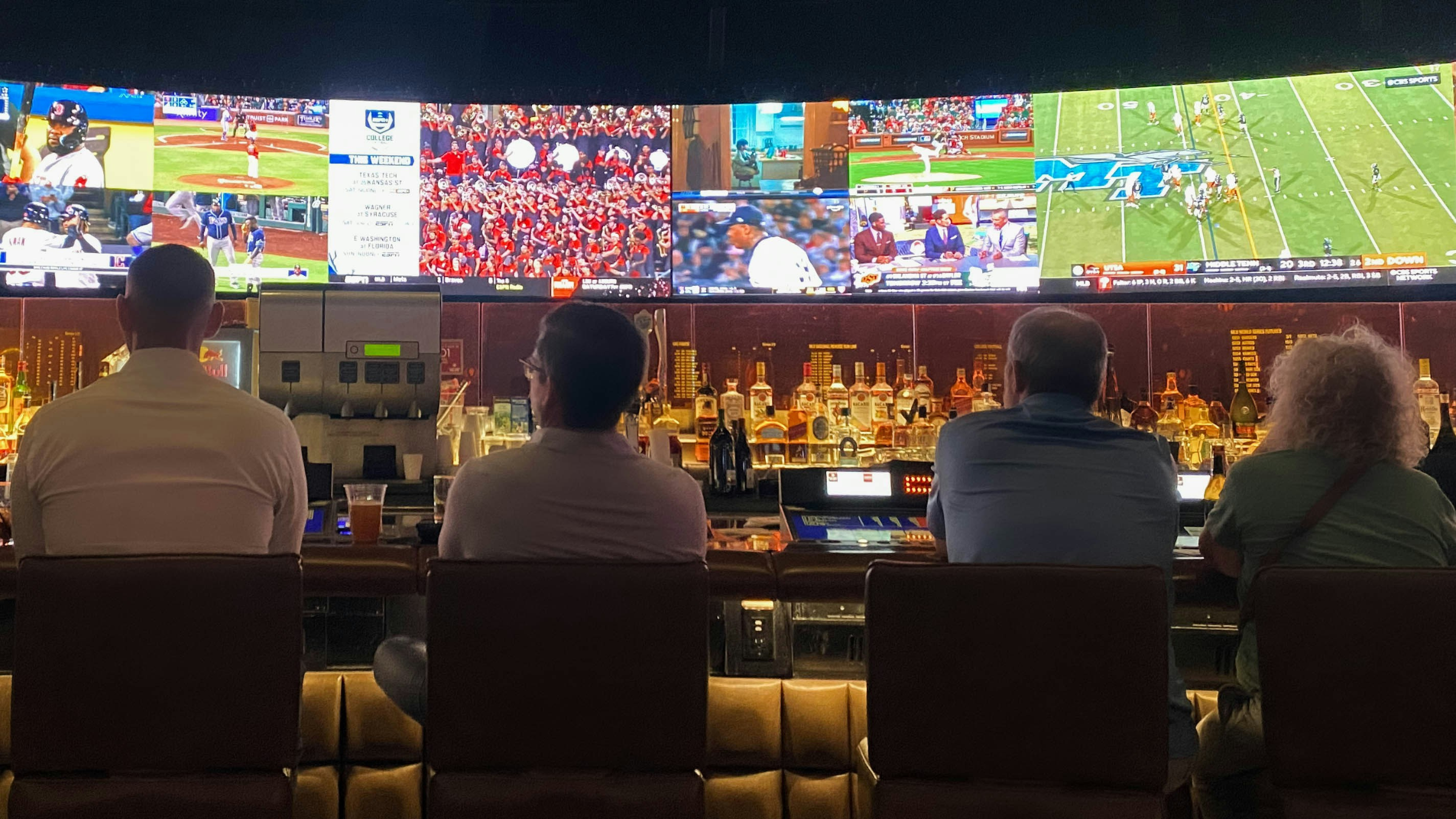 People seated at a sports bar watching multiple live games on large screens above the bar.