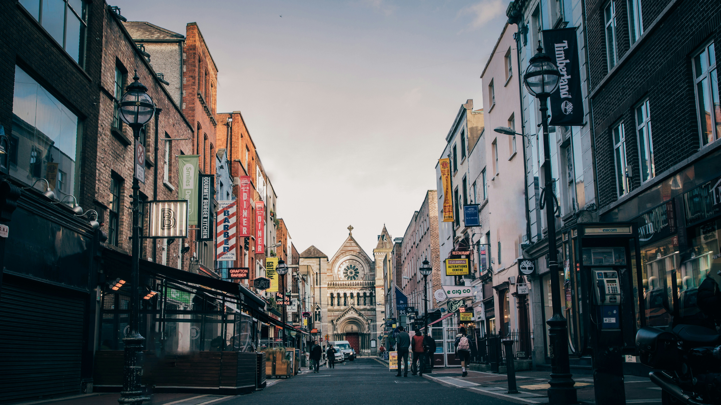 Urban street lined with shops, signs, and pedestrians leading toward a church at the far end.