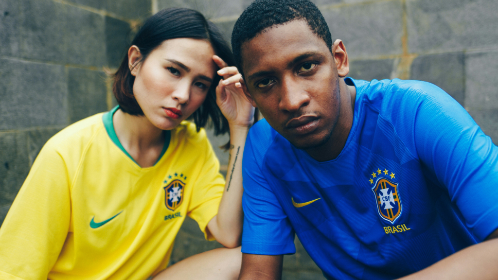 Two people wearing Brazil national team shirts pose against a stone wall background.