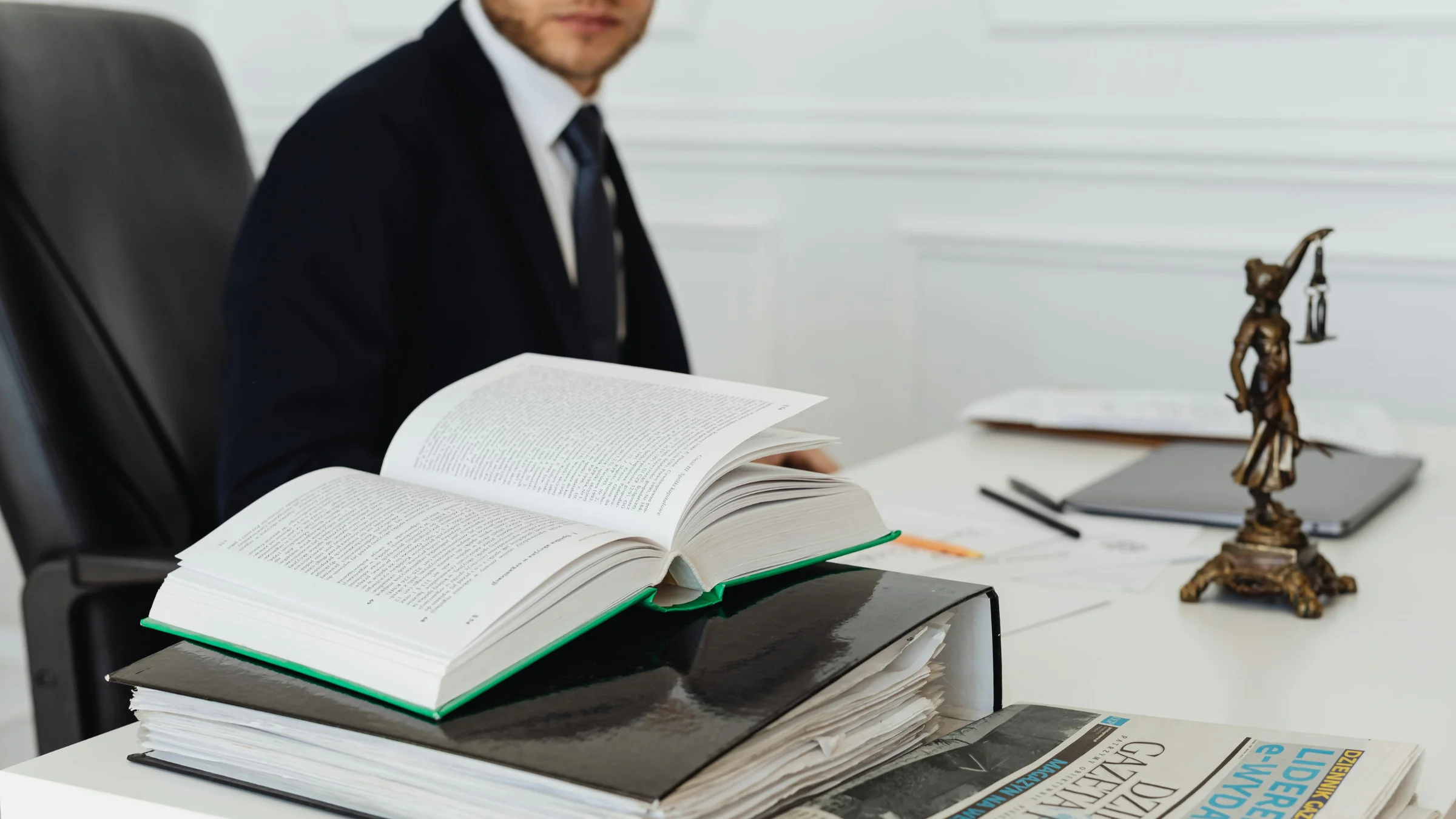 Open law book and legal files on a desk beside a suited lawyer in an office.