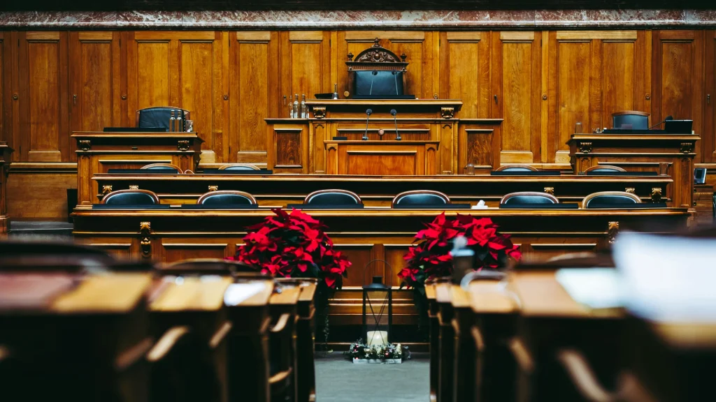 Empty wood-paneled parliamentary or legislative chamber with desks and a central speaker’s chair.