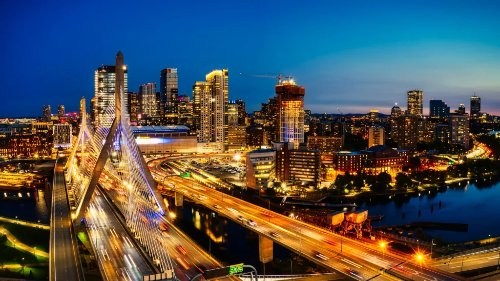 Boston skyline at night with illuminated roads, bridges, and city buildings along the river.
