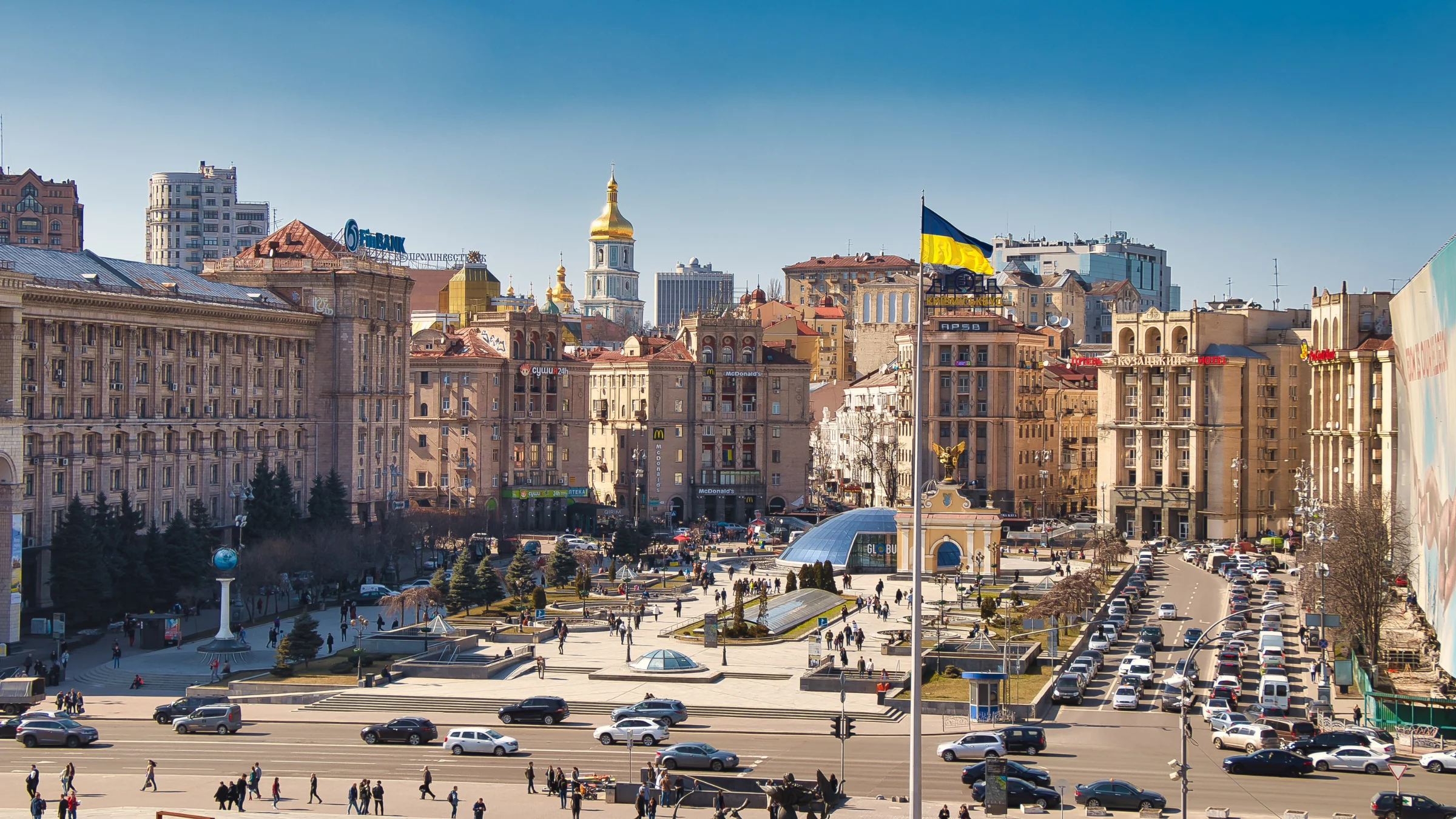 Central Kyiv cityscape with the Ukrainian flag flying above a busy square and surrounding historic buildings.