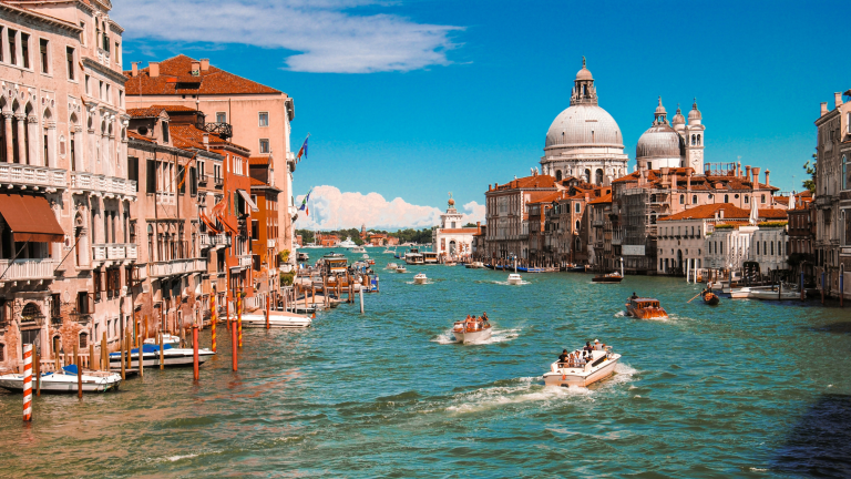 Venice canal scene with boats, historic buildings, and church domes under a clear blue sky.