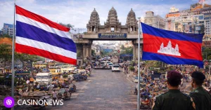 Thai and Cambodian flags at a busy border crossing with an ornate Cambodia gate, traffic, and security personnel visible