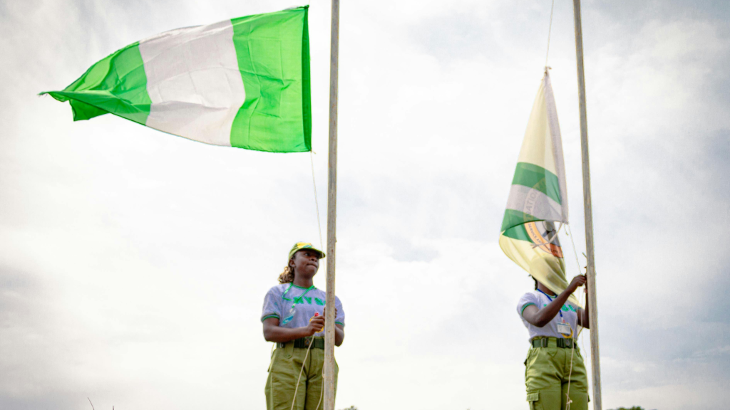 Two individuals raise green and white Nigerian flags on tall poles against a cloudy sky outdoors.