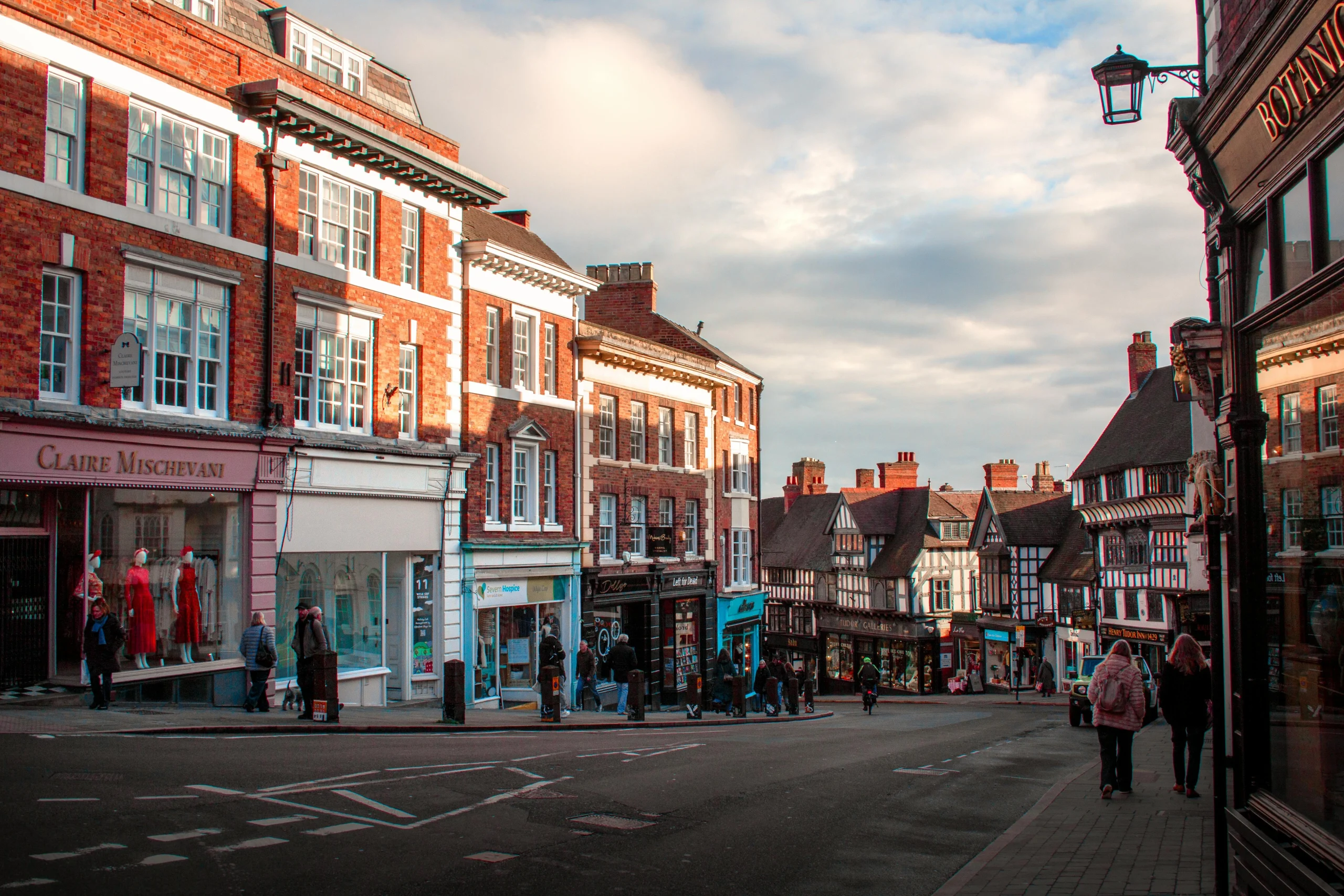 Historic town street with brick buildings, shopfronts, and pedestrians walking along a curved road.