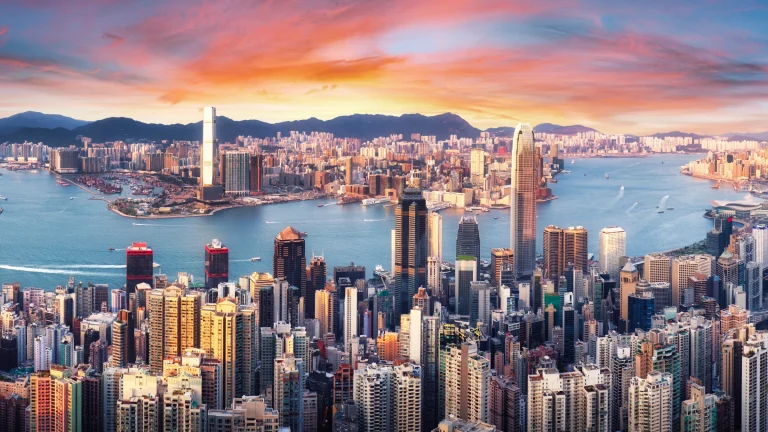Aerial view of Hong Kong skyline and Victoria Harbour at sunset with dense high-rise towers and waterfront traffic.