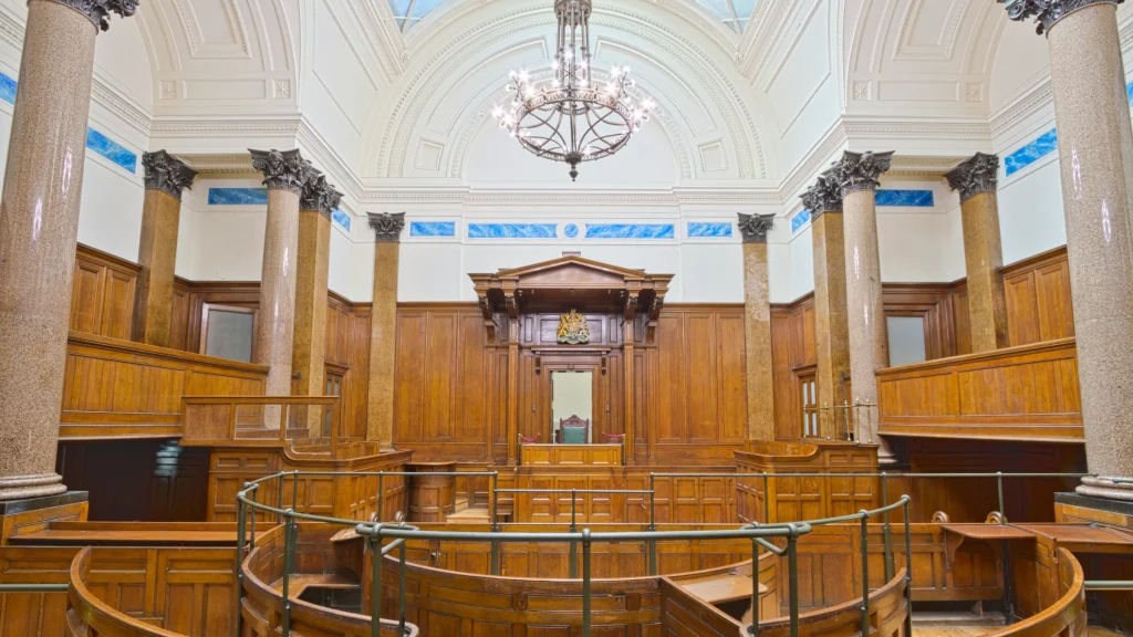 Interior of courtroom with wooden benches, judge’s bench, columns, and chandelier under arched ceiling.