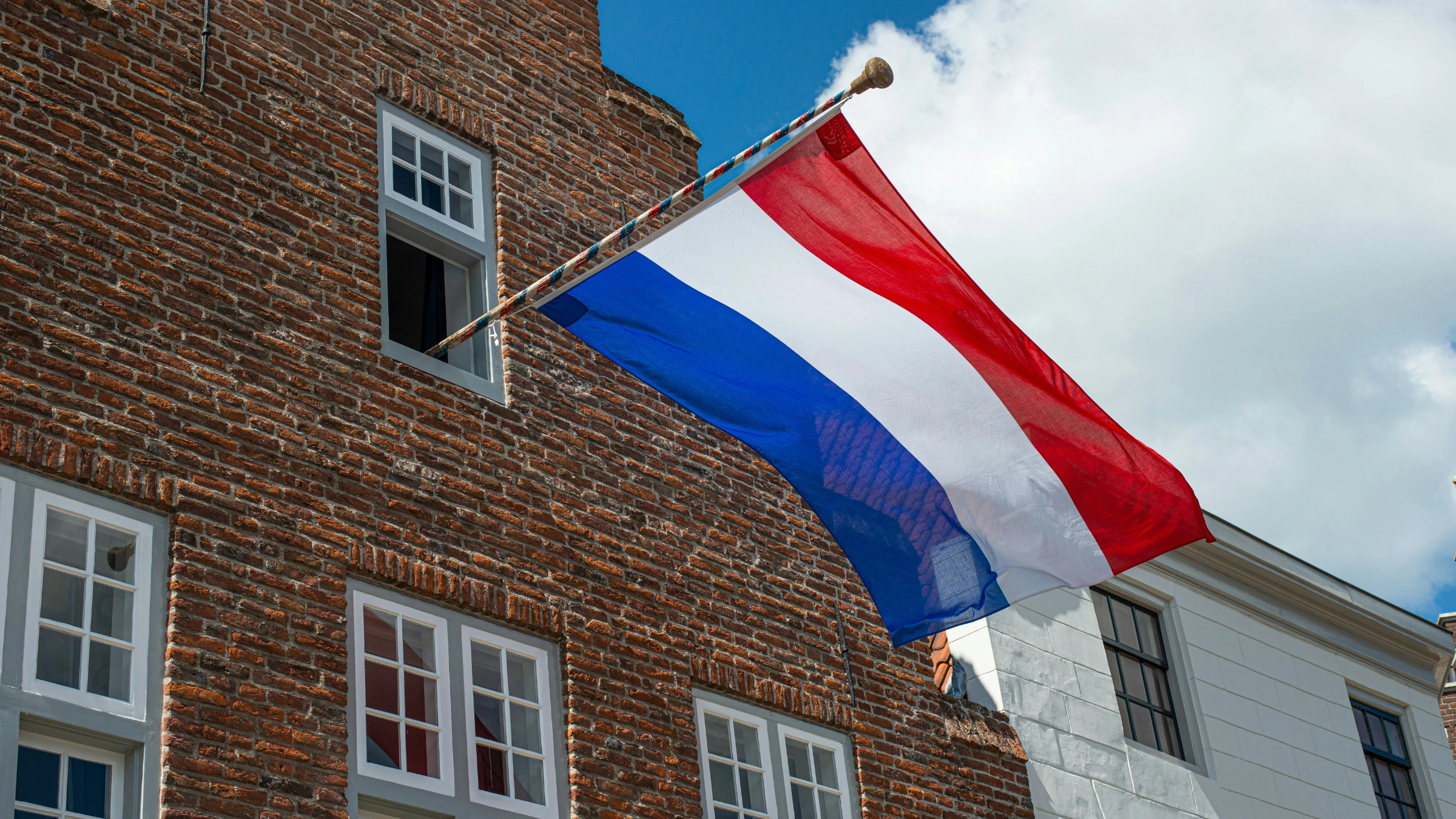 Dutch national flag displayed on historic brick building under partly cloudy sky