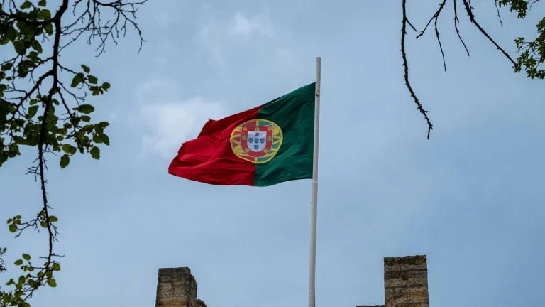 Portuguese national flag flying on a flagpole against a cloudy sky above historic stone walls.