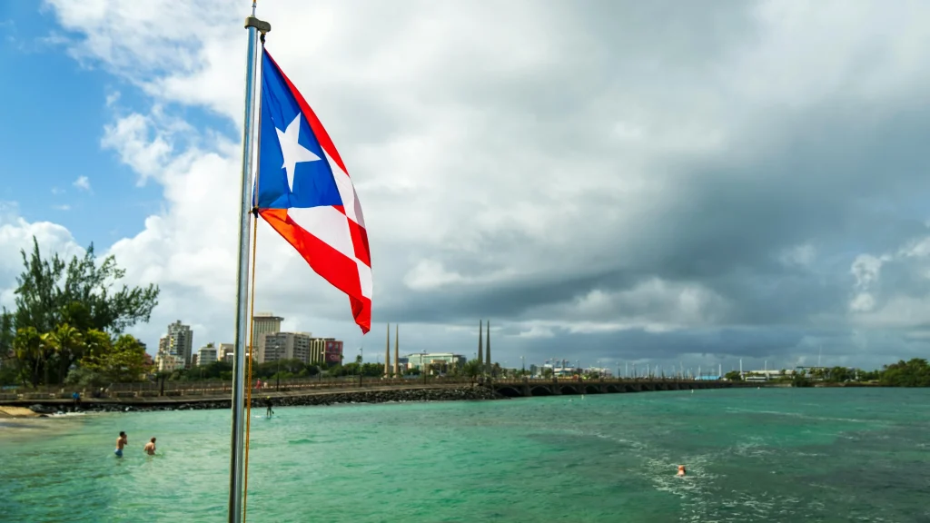 Puerto Rico national flag beside coastal waterfront and city skyline under cloudy skies