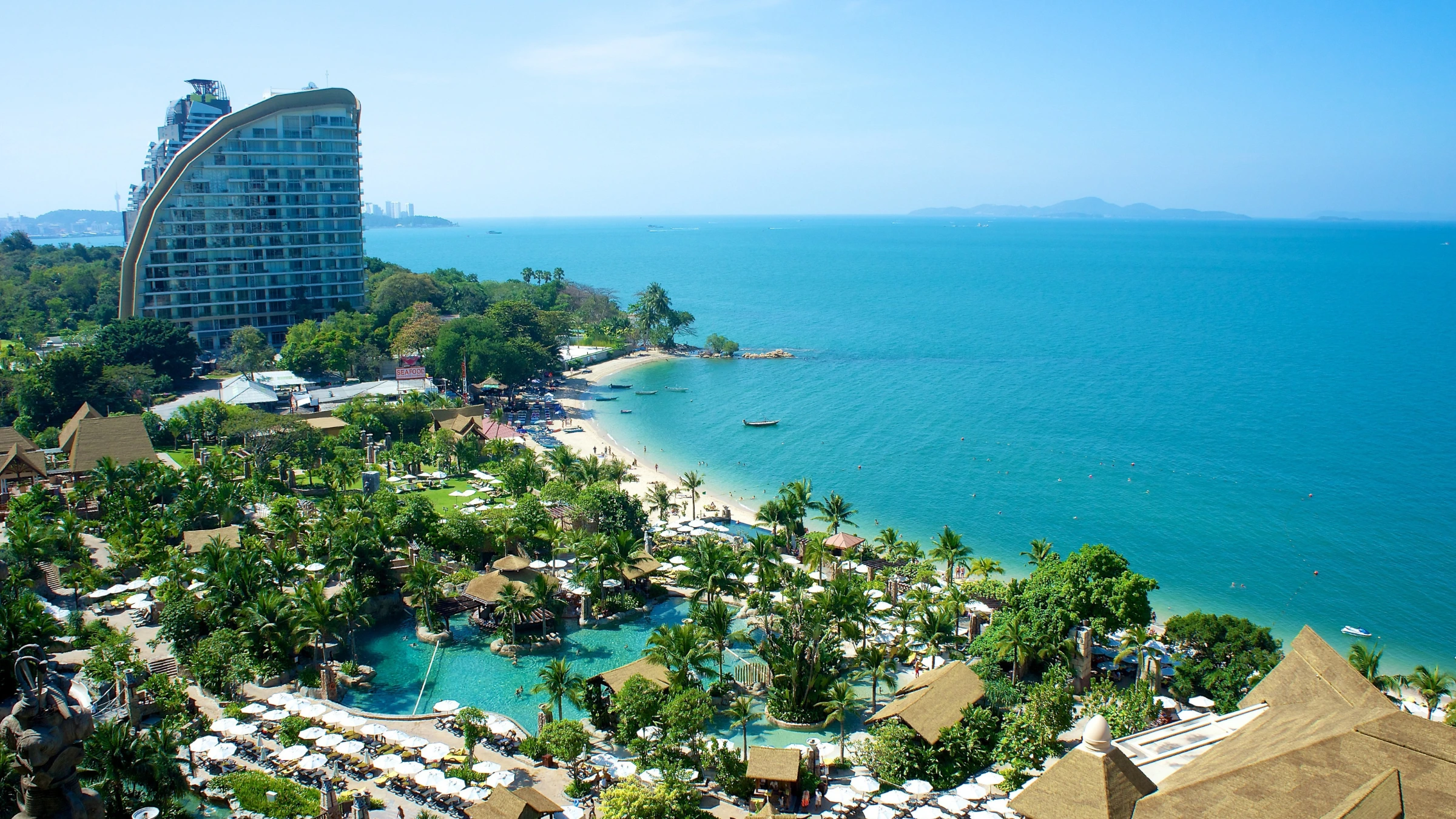 Aerial view of tropical beachfront resort with hotel tower, palm trees, swimming pools, and turquoise sea.
