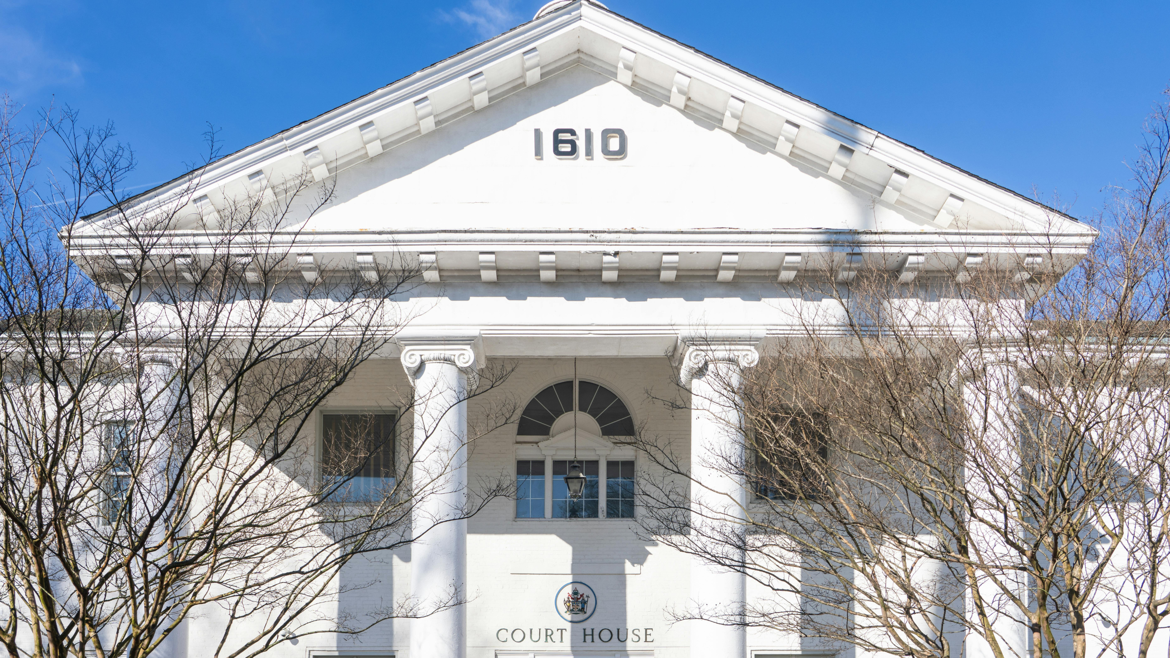 White courthouse building with columns and “1610” inscription above entrance under clear blue sky.